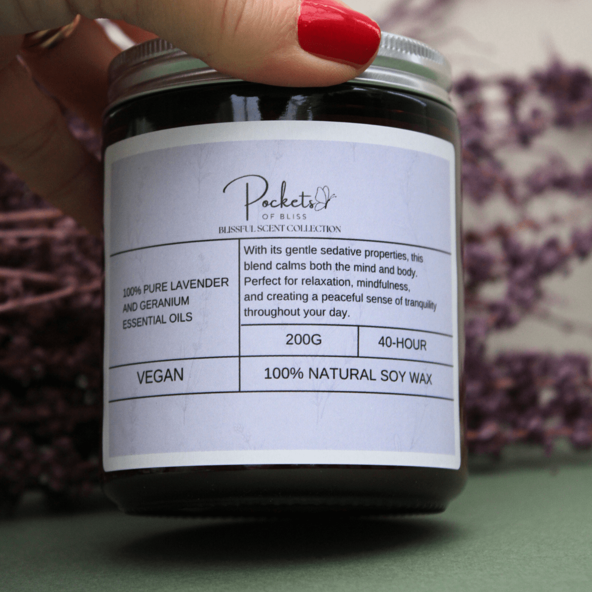 Sian founder of pockets of bliss holding Lavender & Geranium Soy Candle, with red nail varnish and dry lavender in the background. - Pockets of Bliss
